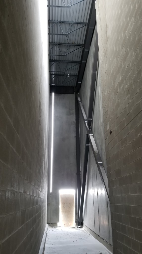 An image of a narrow indoor passageway made of concrete with a metal roof and exposed beams demonstrating a daylighting de...