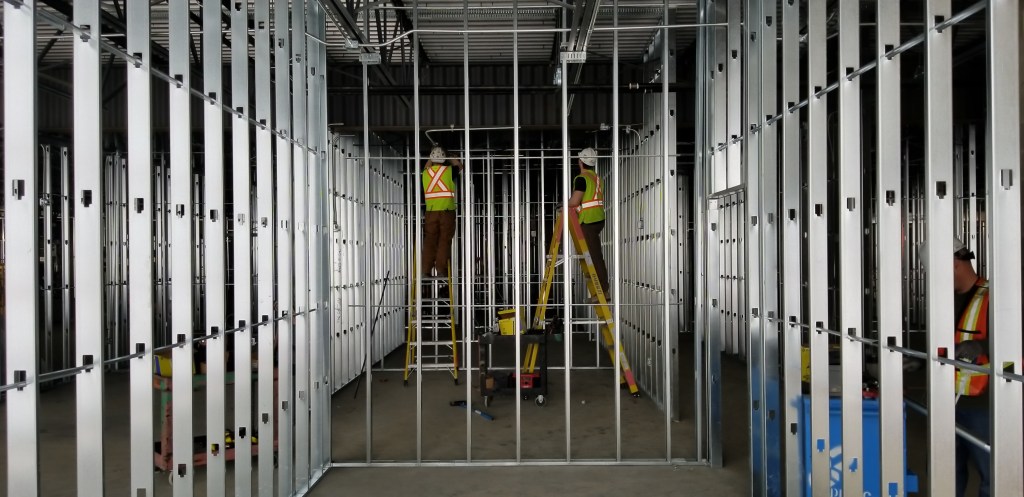 Three workers are constructing metal framing for interior walls in an industrial building.