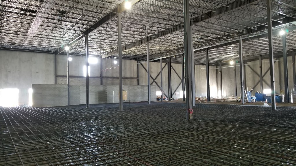 A large warehouse with exposed steel beams and pipes, and black metal grating covering the floor.
