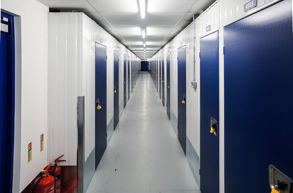 The long, well-lit corridor is lined with rows of closed storage units, featuring secure doors with yellow padlocks and fi...