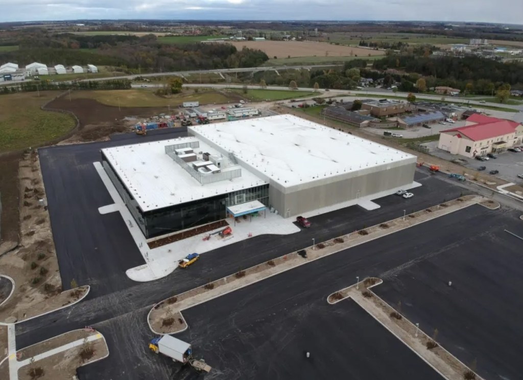 An aerial view of a large industrial building and parking lot set amidst several smaller buildings.