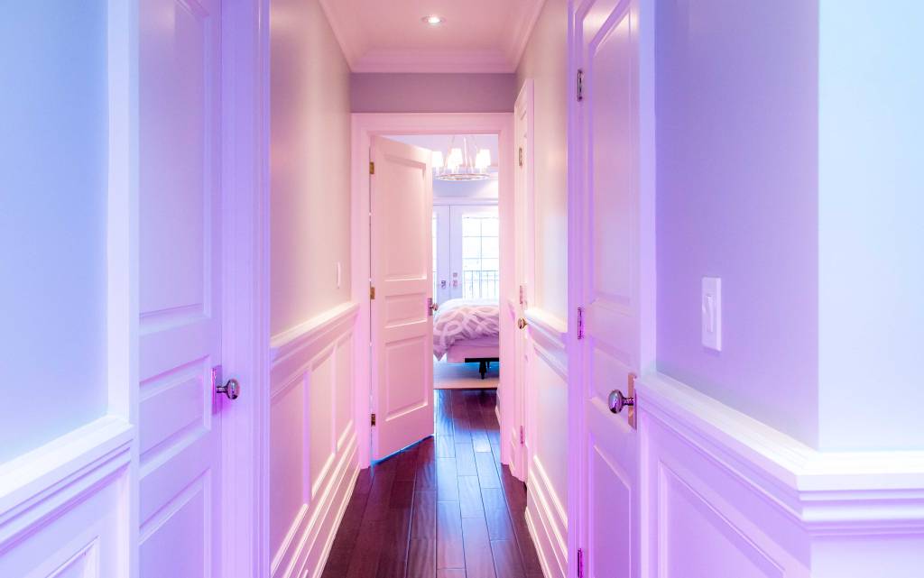 Unmade bed in a narrow hallway with white walls, and doors leading to not-Seen roomschts. Crisp white curtains, purple hues.
